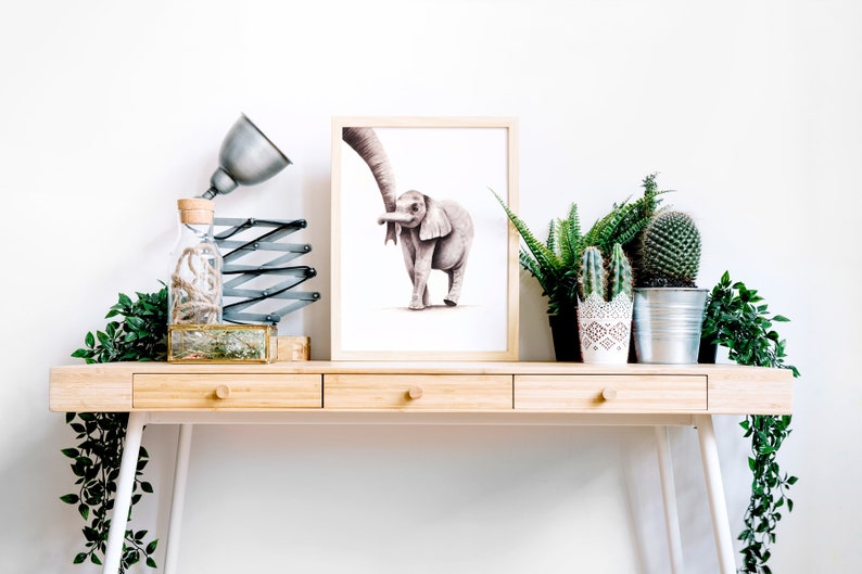 May include: A framed watercolor painting of an elephant and a baby elephant walking together, displayed on a wooden desk with a lamp, a glass bottle, and potted plants.