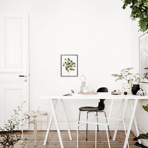May include: A white table with a white chair and a black chair. There is a framed print of a green plant on the wall behind the table. There are plants in pots on the table and on the floor.