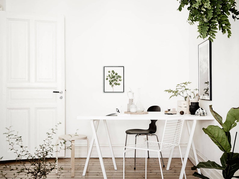 May include: A white table with a white chair and a black chair. There is a framed print of a green plant on the wall behind the table. There are plants in pots on the table and on the floor.