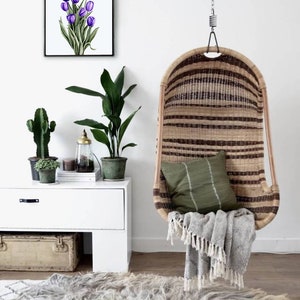 May include: A white wall with a framed print of purple tulips, a white dresser with a potted plant and a cactus, a hanging wicker chair with a green cushion and a white blanket, and a fluffy gray rug on the floor.
