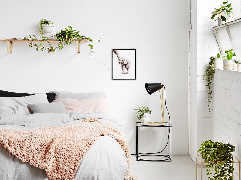 May include: A framed watercolor painting of a baby elephant with its trunk raised in the air. The painting is hanging on a white wall in a bedroom setting with a bed, nightstand, and plants.