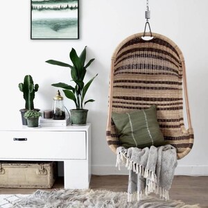 K&ouml;nnte beinhalten: Ein h&auml;ngender Korbstuhl mit einem gr&uuml;nen Kissen und einer grauen Decke. Der Stuhl h&auml;ngt an einer Kette und steht vor einer wei&szlig;en Wand mit einem gerahmten Druck eines gr&uuml;nen Landschaftsbildes.
