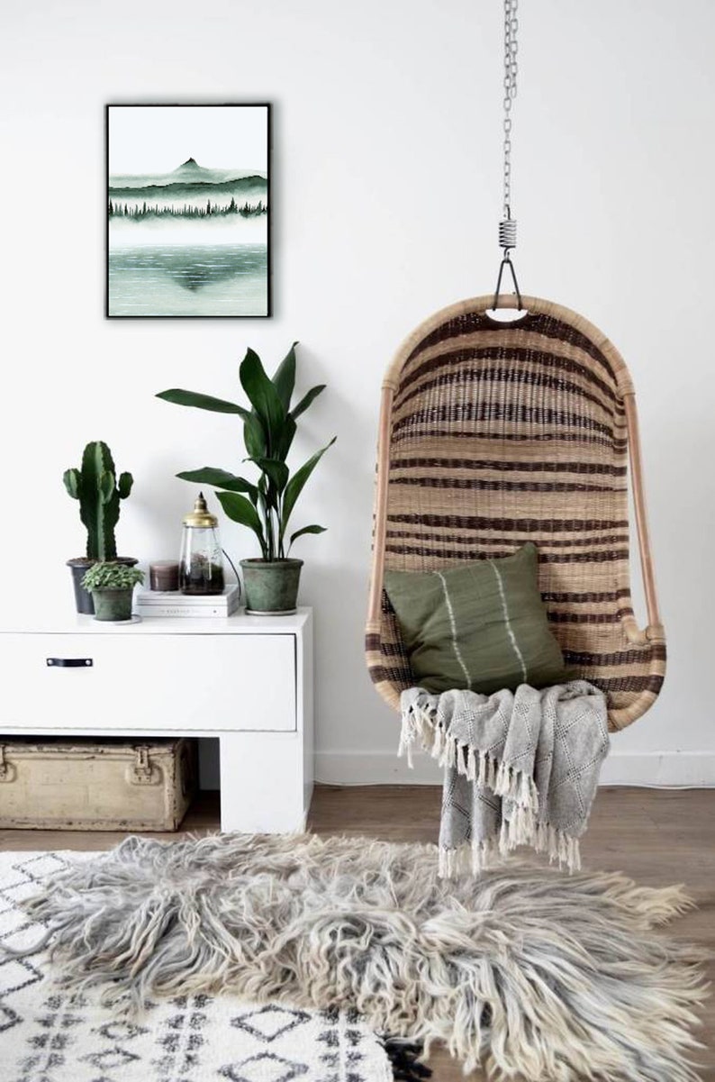 K&ouml;nnte beinhalten: Ein h&auml;ngender Korbstuhl mit einem gr&uuml;nen Kissen und einer grauen Decke. Der Stuhl h&auml;ngt an einer Kette und steht vor einer wei&szlig;en Wand mit einem gerahmten Druck eines gr&uuml;nen Landschaftsbildes.