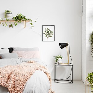 May include: A white bedroom with a bed, a nightstand, and a lamp. The bed has a white duvet and a pink throw blanket. The nightstand is black and has a gold lamp on it. There is a framed print of a green plant on the wall.