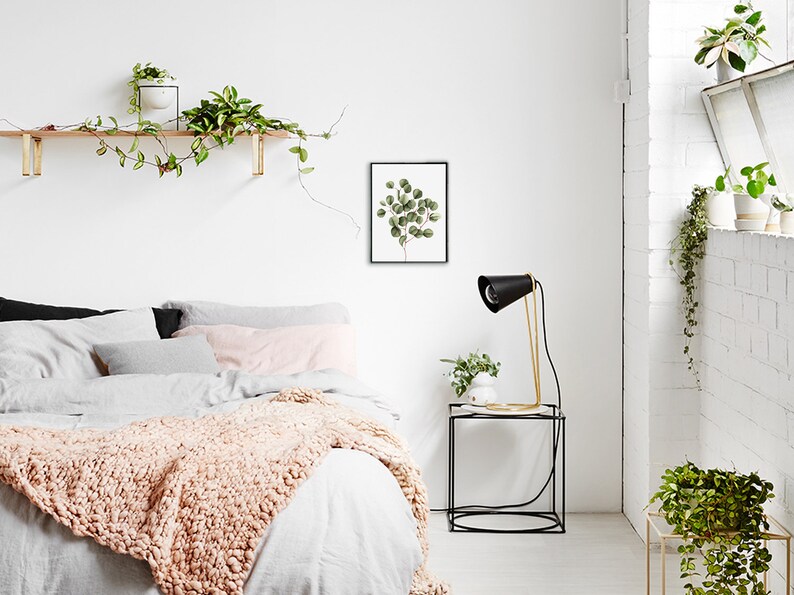 May include: A white bedroom with a bed, a nightstand, and a lamp. The bed has a white duvet and a pink throw blanket. The nightstand is black and has a gold lamp on it. There is a framed print of a green plant on the wall.