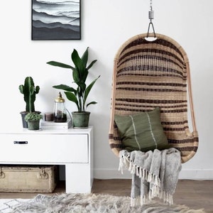 May include: A hanging wicker chair with a green cushion and a gray throw blanket. The chair is hanging from a white ceiling and there is a rug on the floor. There is a white cabinet with a cactus and a plant on top of it.