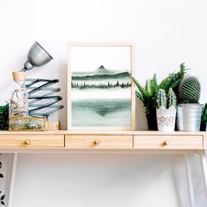 K&ouml;nnte beinhalten: Ein gerahmtes Aquarellbild einer Bergkette mit einem See im Vordergrund. Das Bild befindet sich auf einem Holztisch mit einer Lampe, einer Glasflasche und mehreren Topfpflanzen.