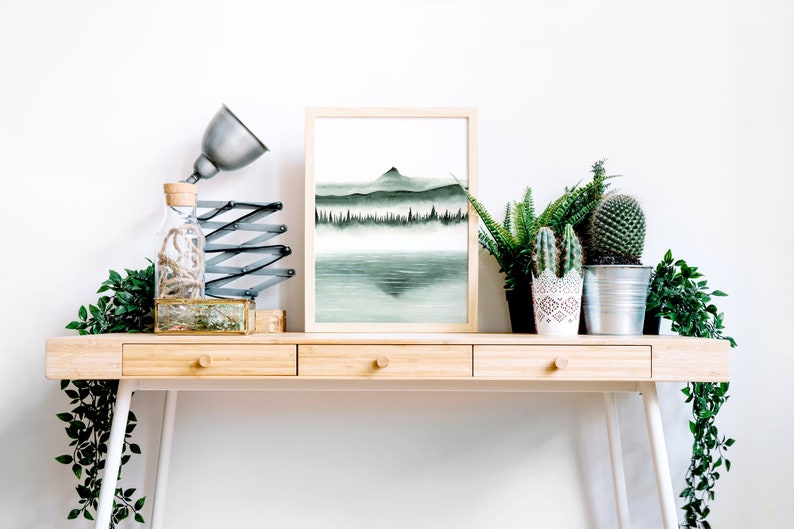 K&ouml;nnte beinhalten: Ein gerahmtes Aquarellbild einer Bergkette mit einem See im Vordergrund. Das Bild befindet sich auf einem Holztisch mit einer Lampe, einer Glasflasche und mehreren Topfpflanzen.