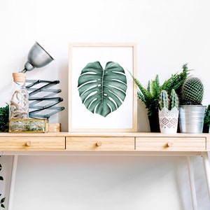 May include: A wooden desk with three drawers and white legs. A framed print of a green monstera leaf hangs above the desk. There are several potted plants on the desk, including a cactus, a fern, and a succulent.
