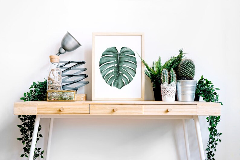 May include: A wooden desk with three drawers and white legs. A framed print of a green monstera leaf hangs above the desk. There are several potted plants on the desk, including a cactus, a fern, and a succulent.