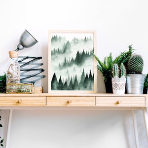 May include: A framed watercolor print of a misty forest scene in shades of green, displayed on a wooden desk with potted plants and a lamp.
