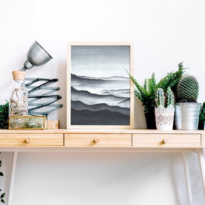 May include: A wooden desk with three drawers and white legs. A framed black and white print of a mountain range hangs above the desk. A lamp with a silver shade and a glass bottle with a cork top are on the desk. There are three potted plants on the desk, including a cactus and a fern.