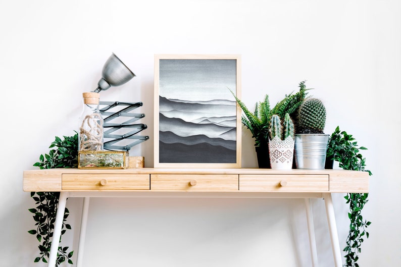 May include: A wooden desk with three drawers and white legs. A framed black and white print of a mountain range hangs above the desk. A lamp with a silver shade and a glass bottle with a cork top are on the desk. There are three potted plants on the desk, including a cactus and a fern.