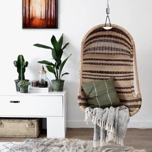May include: A hanging wicker chair with a green cushion and a grey throw blanket. The chair is hanging from a white ceiling with a chain. There is a rug on the floor and a white dresser with plants and a painting of a forest scene on the wall.