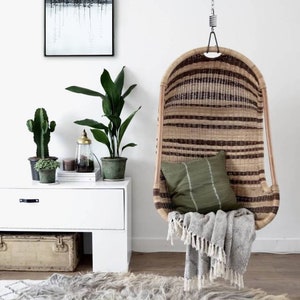 Pu&ograve; includere: Una sedia a dondolo in vimini con un cuscino verde e una coperta grigia. La sedia &egrave; appesa a un soffitto bianco con una catena. C'&egrave; una cassettiera bianca con un cactus e una pianta sopra. C'&egrave; un tappeto sul pavimento con un motivo bianco e nero.