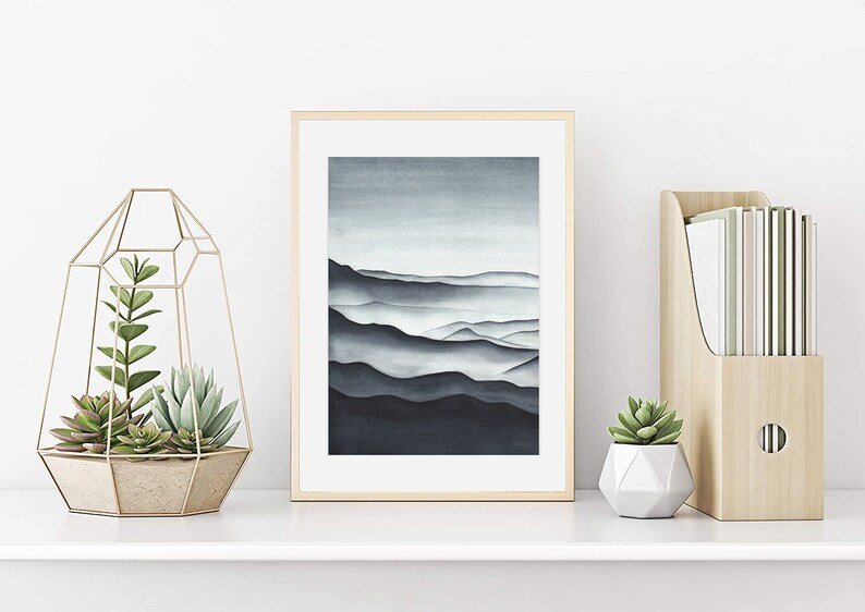 May include: A framed watercolor painting of a mountain range in shades of blue and gray. The painting is displayed on a white shelf with a gold geometric terrarium and a wooden magazine holder.