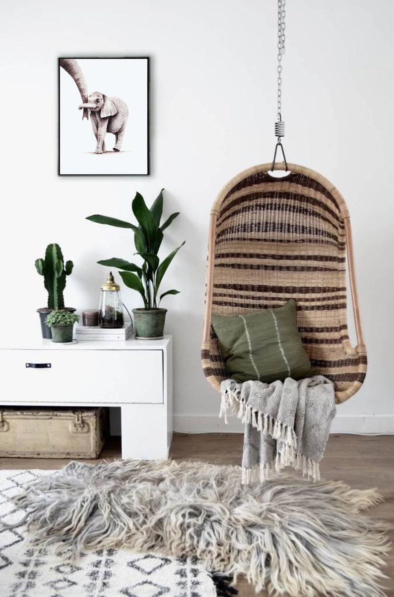 May include: A hanging wicker chair with a green cushion and a gray throw blanket. The chair is hanging from a chain and is in front of a white wall. There is a white dresser with a framed print of an elephant and a potted plant on top of it. A gray and white rug is on the floor.