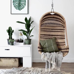 May include: A hanging wicker chair with a green cushion and a white blanket. The chair is hanging from a chain and is in front of a white wall with a framed print of a green leaf.