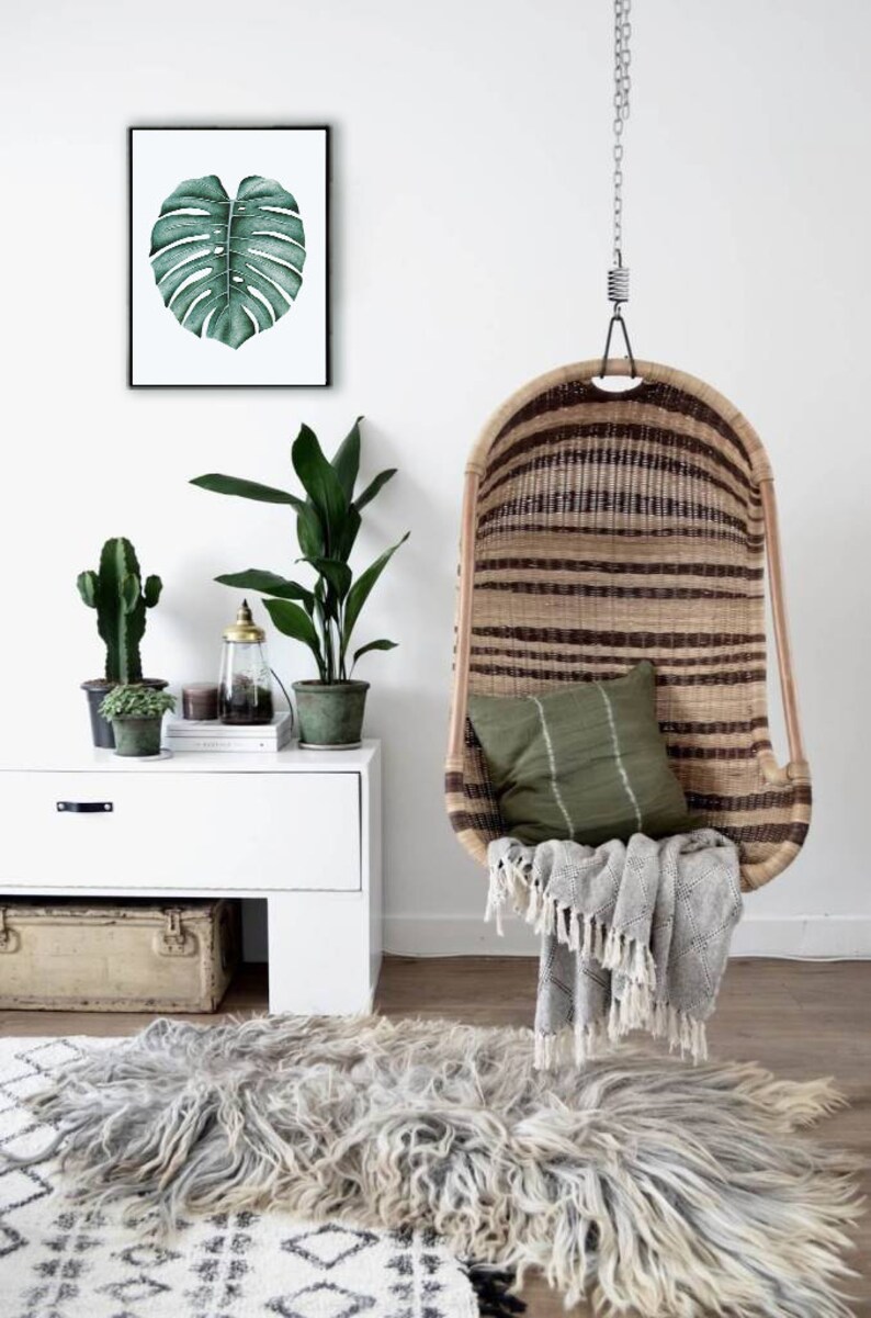May include: A hanging wicker chair with a green cushion and a white blanket. The chair is hanging from a chain and is in front of a white wall with a framed print of a green leaf.