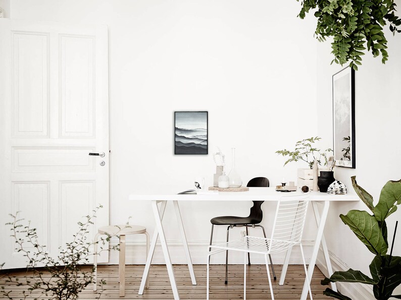 May include: A white table with a white trestle base and two chairs. One chair is black and the other is white. There is a framed black and white print on the wall behind the table. There are plants in the room.