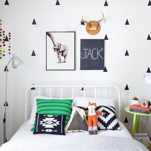 May include: A white bed with a green and white striped pillow, a black and white geometric pillow, a gray pillow with a mountain design, and a stuffed fox wearing a tie. The bed is in a room with white walls and black triangle decals. The name "JACK" is written on a black chalkboard hanging on the wall.
