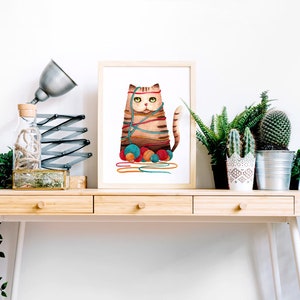 Puede incluir: Una ilustraci&oacute;n enmarcada de un gato marr&oacute;n y naranja enredado en hilo de colores con ovillos de hilo a sus pies. La ilustraci&oacute;n est&aacute; sobre un escritorio de madera con una l&aacute;mpara, una botella de vidrio y plantas en macetas.