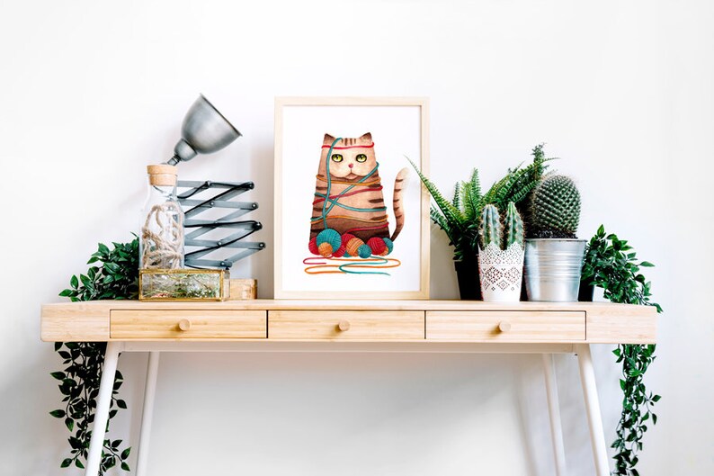 Puede incluir: Una ilustraci&oacute;n enmarcada de un gato marr&oacute;n y naranja enredado en hilo de colores con ovillos de hilo a sus pies. La ilustraci&oacute;n est&aacute; sobre un escritorio de madera con una l&aacute;mpara, una botella de vidrio y plantas en macetas.