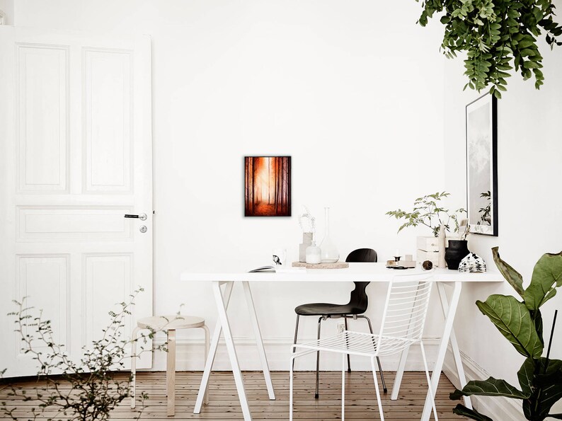 May include: A white table with a white chair and a black chair. A framed painting of a forest scene hangs on the wall behind the table. There are plants on the table and on the floor.