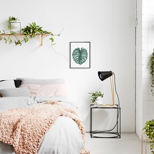 May include: A white bedroom with a bed, a nightstand, and a framed print of a green leaf. The bed has a white duvet and a pink throw blanket. The nightstand has a black lamp with a gold base and a small plant. The print is on a white background and has a black frame.