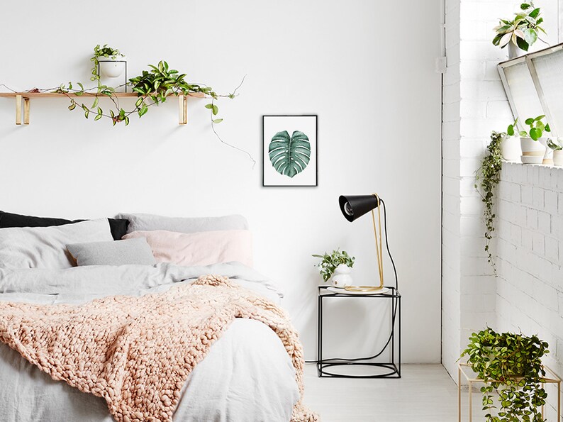 May include: A white bedroom with a bed, a nightstand, and a framed print of a green leaf. The bed has a white duvet and a pink throw blanket. The nightstand has a black lamp with a gold base and a small plant. The print is on a white background and has a black frame.