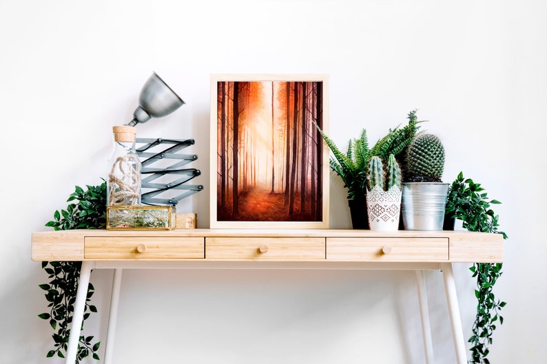 May include: A framed painting depicting a forest path with tall trees and a warm, orange glow. The painting is displayed on a wooden desk with a lamp, a glass bottle, and potted plants.
