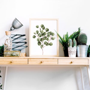 May include: A framed print of a watercolor painting of a eucalyptus branch with green leaves on a white background. The print is sitting on a wooden desk with a lamp, a glass bottle, and several potted plants.