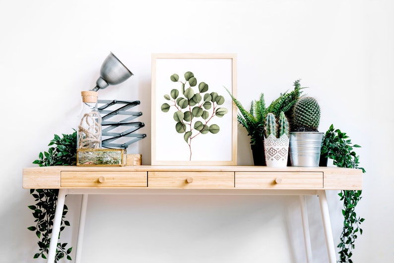 May include: A framed print of a watercolor painting of a eucalyptus branch with green leaves on a white background. The print is sitting on a wooden desk with a lamp, a glass bottle, and several potted plants.