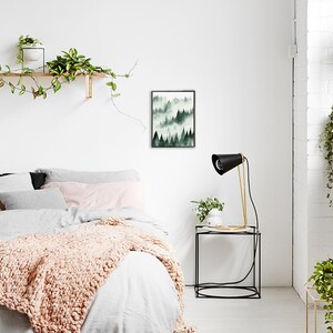 May include: A bedroom with a white wall, a bed with a white duvet and a pink throw blanket, a black framed print of a watercolor painting of a forest, a black lamp on a black side table, and a potted plant on the table.
