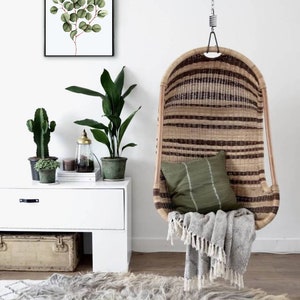 May include: A hanging wicker chair with a green cushion and a gray throw blanket. The chair is hanging from a white ceiling with a chain. There is a white dresser with a green plant and a cactus on top of it. There is a rug on the floor with a black and white pattern.