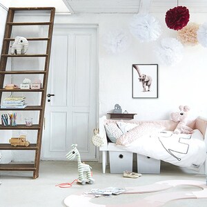 May include: A white and pink nursery with a wooden ladder shelf, a white crib with pink bedding, a stuffed elephant toy, and a wooden giraffe toy. The room is decorated with white pom-poms and a framed print of an elephant.