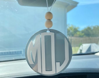 May include: A gray and white car air freshener with the monogram "MOJ" hanging from the rearview mirror. The air freshener is attached to a string with two wooden beads.