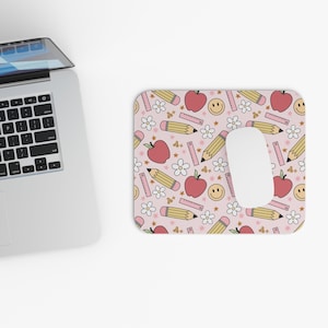 May include: A rectangular mousepad with a light pink background and a repeating pattern of red apples, yellow pencils, white flowers, rulers, stars, and smiling faces. The mousepad has a cutout for the mouse.