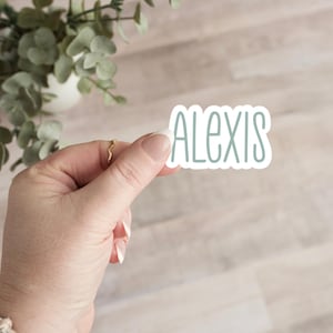 May include: A sticker with the name "ALEXIS" in a pale blue font. The sticker is held by a hand with a gold ring. The background is a light wood-grain pattern with a plant in the upper left corner.