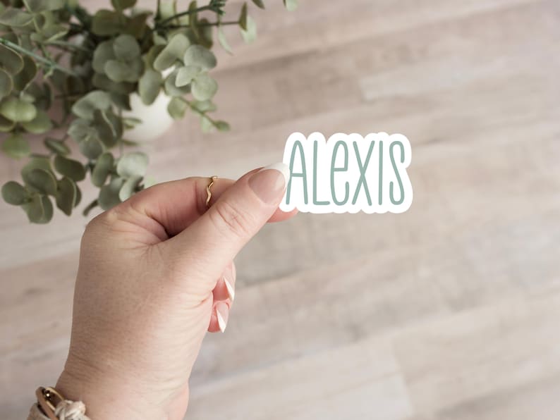 May include: A sticker with the name "ALEXIS" in a pale blue font. The sticker is held by a hand with a gold ring. The background is a light wood-grain pattern with a plant in the upper left corner.