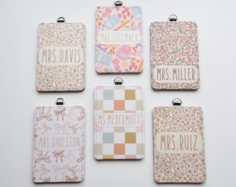 May include: Six rectangular ID badge holders with metal loops, each featuring unique patterns including floral prints, bows, school supplies, and a checkered design. Each holder displays a personalized name label: Mrs. Davis, Mrs. Stelmack, Mrs. Miller, Mrs. Gholston, Ms. McDermott, and Mrs. Ruiz.