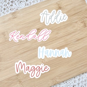 May include: Four name stickers: "Addie" in grey, "Kendall" in pink, "Hannah" in light blue, and "Maggie" in mauve. The stickers are on a light brown wooden cutting board, with a white textured background.