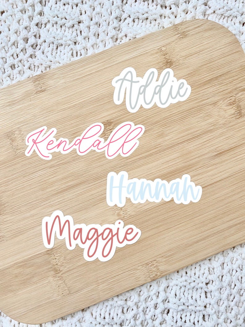 May include: Four name stickers: "Addie" in grey, "Kendall" in pink, "Hannah" in light blue, and "Maggie" in mauve. The stickers are on a light brown wooden cutting board, with a white textured background.