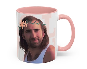 May include: A white ceramic mug with a pink rim and handle. The mug features a photo of Nicolas Cage with a flower crown on his head. The text "Not" is visible on the mug.