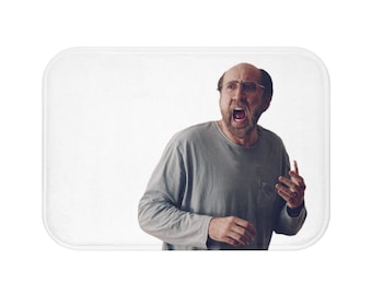 May include: A bath mat featuring a photo of a man with a surprised expression, wearing glasses and a grey shirt. He is yelling with his mouth open.