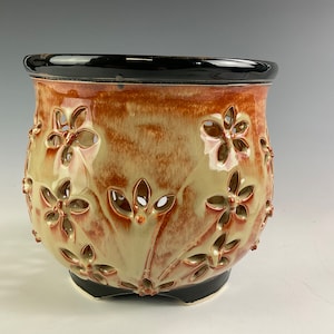 May include: A ceramic planter with a black rim and intricate floral cutouts. The planter is glazed in a light beige with brown accents.