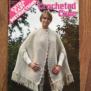 May include: A vintage crochet pattern leaflet for a crocheted cape. The leaflet features a woman wearing the cape, which is a light beige color with fringe trim. The leaflet is titled "Crocheted Cape" and has the text "Extra Easy Instructions" in pink.