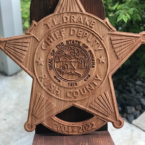 May include: A wooden star-shaped plaque with the text "T.L. DRAKE CHIEF DEPUTY" and "SEAL OF THE STATE OF INDIANA" engraved on it. The plaque also includes "RUSH COUNTY" and the years "2004-2022". The plaque is mounted on a dark brown wooden stand.