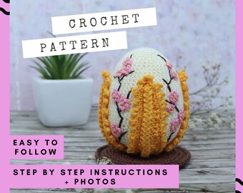 Op de afbeelding: Een gehaakte eivormige decoratie met een crèmekleurig lichaam, versierd met roze bloemenborduurwerk en een gele, getextureerde rand. De tekst "CROCHET PATTERN" staat boven het ei. De afbeelding bevat ook de tekst "EASY TO FOLLOW" en "STEP BY STEP INSTRUCTIONS + PHOTOS".