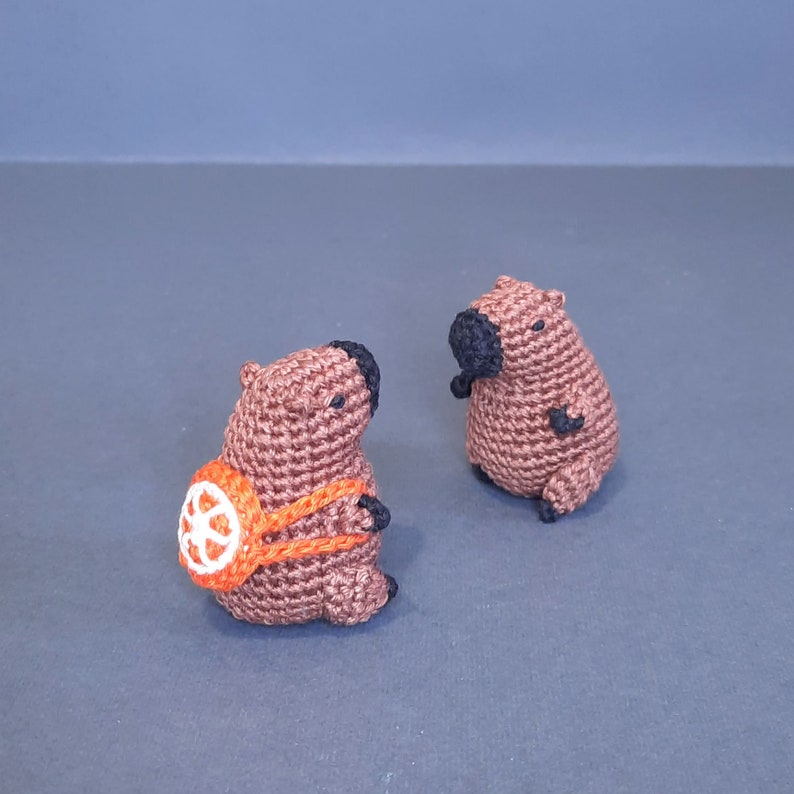 May include: Two brown crocheted capybara plushies. One capybara is wearing an orange backpack with a white wheel design.